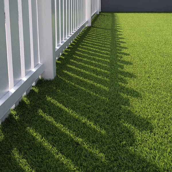 Sunlight and shadow of white wooden fence on green artificial turf surface in front yard of home, selective focus with high angle view