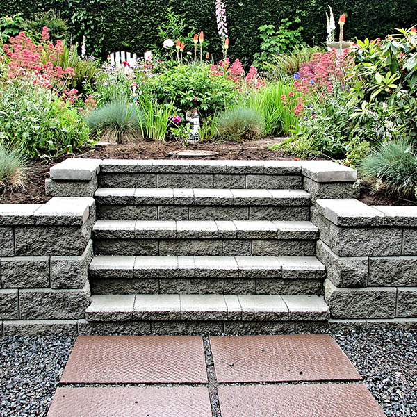 Block retaining wall with incorporated staircase into existing garden landscape consisting of perennial flowers and bushes with stepping stones leading up to the steps.