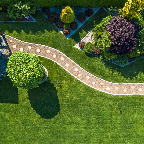 Aerial view of a vibrant garden featuring a winding stone path, ornamental shrubs, and well-manicured grass under bright sunlight, creating a serene atmosphere.